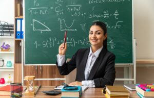 impressed-young-female-teacher-sits-table-with-school-tools-raising-pen-classroom_141793-106366
