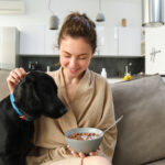 Cosy mornings with best friend. Young woman eating cereals for breakfast, sitting with her dog on sofa and smiling, starting her morning with proper meal
