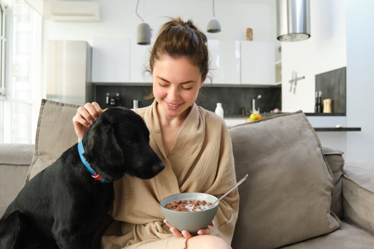 Cosy mornings with best friend. Young woman eating cereals for breakfast, sitting with her dog on sofa and smiling, starting her morning with proper meal