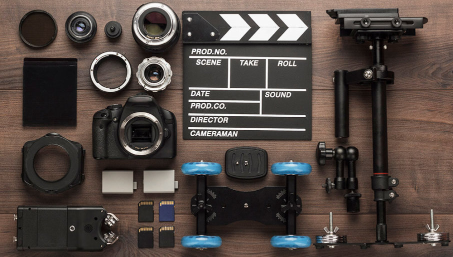 Flat lay of filmmaking equipment with camera gear, lenses, clapperboard, memory cards, and stabilizer on a wooden surface.