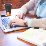 A close-up of a student’s hands typing on a laptop next to a notebook and tea, representing personalized academic writing assistance for research and essay composition.