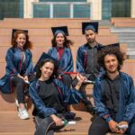 Five diverse graduates in blue caps and gowns sitting on outdoor steps holding diplomas, representing academic success and the value of Ethical Assignment Help for University Students.