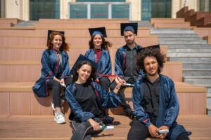 Five diverse graduates in blue caps and gowns sitting on outdoor steps holding diplomas, representing academic success and the value of Ethical Assignment Help for University Students.