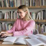 A female student smiling while typing on a laptop in a library, researching how to write an assignment for university UK.