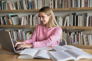 A female student smiling while typing on a laptop in a library, researching how to write an assignment for university UK.