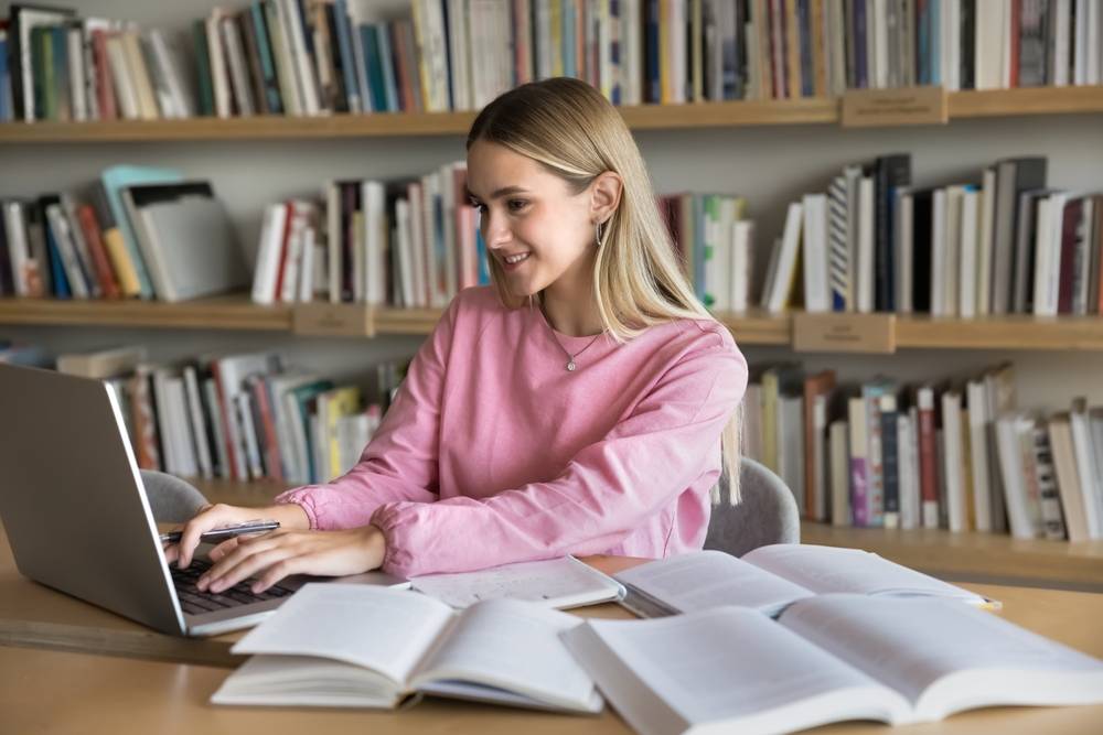 A female student smiling while typing on a laptop in a library, researching how to write an assignment for university UK.