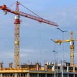 Three tall tower cranes and scaffolding at a busy job site managed by a Construction Company London, set against a blue sky.