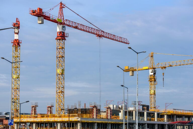 Three tall tower cranes and scaffolding at a busy job site managed by a Construction Company London, set against a blue sky.