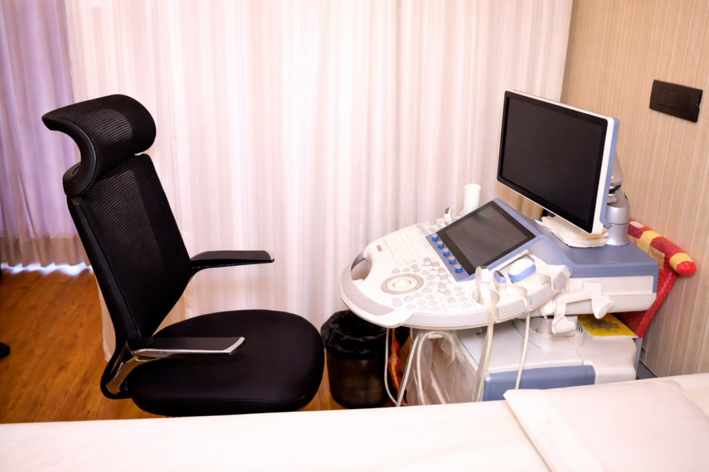 Modern ultrasound examination room with a diagnostic imaging machine, adjustable chair, monitor, and patient bed in a clean clinical setting