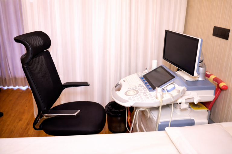 Modern ultrasound examination room with a diagnostic imaging machine, adjustable chair, monitor, and patient bed in a clean clinical setting