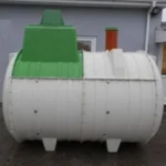White cylindrical tank with green top section and a red vertical pipe, positioned outdoors beside a building.
