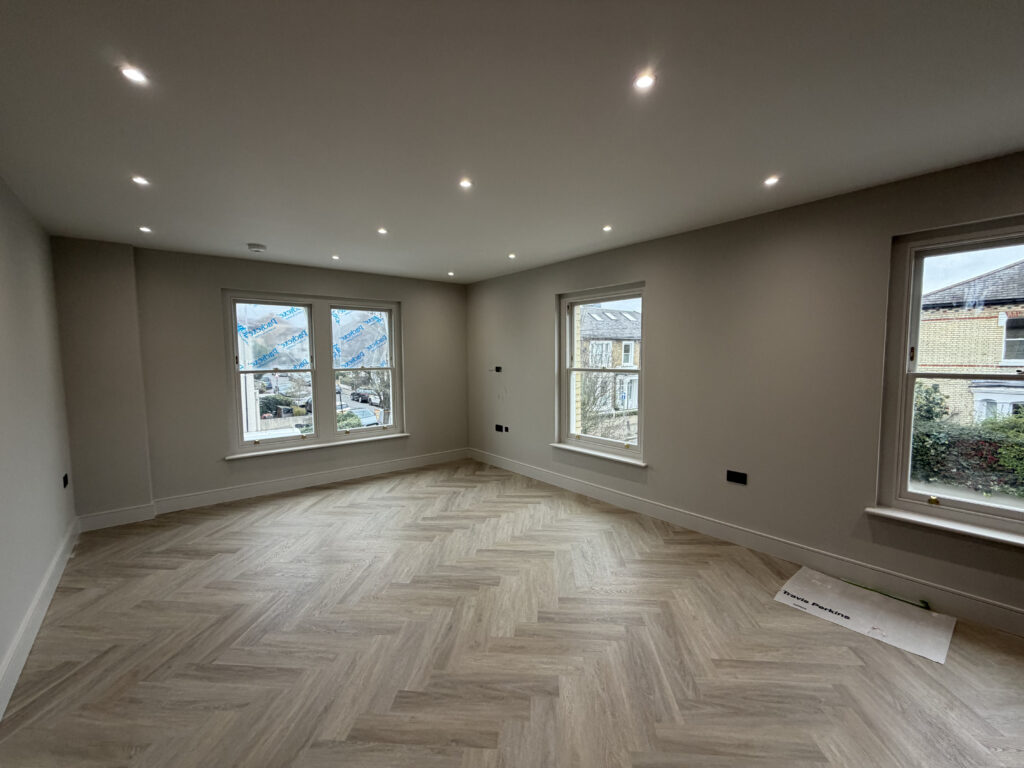 A newly renovated, spacious room in a London property featuring light-colored herringbone wood flooring, neutral grey walls, and modern recessed spotlighting—showcasing a high-standard of home improvement London.