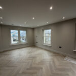 A newly renovated, spacious room in a London property featuring light-colored herringbone wood flooring, neutral grey walls, and modern recessed spotlighting—showcasing a high-standard of home improvement London.