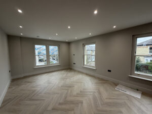 A newly renovated, spacious room in a London property featuring light-colored herringbone wood flooring, neutral grey walls, and modern recessed spotlighting—showcasing a high-standard of home improvement London.