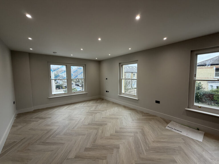 A newly renovated, spacious room in a London property featuring light-colored herringbone wood flooring, neutral grey walls, and modern recessed spotlighting—showcasing a high-standard of home improvement London.