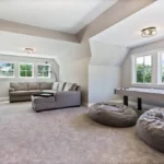 A spacious, carpeted multi-purpose room created through a garage conversion, featuring a large sectional sofa, bean bags, and an air hockey table under a vaulted ceiling.
