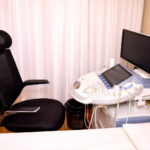 Modern ultrasound machine with monitor and probes beside an adjustable black chair in a clean clinical examination room.