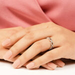 Elegant infinity twist diamond ring with pavé-set stones worn on a hand against a soft pink background.