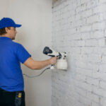 man painting old brick wall in white color with paint sprayer