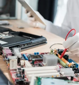 Technician testing a computer motherboard with a multimeter probe during laptop repair and electronic hardware diagnostics.
